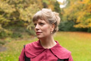 Photo of Sarah Perry. Sarah is standing outside in a lawned area with trees. Sarah is wearing a red top and is looking away from the camera.