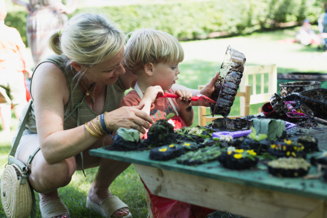 Photo of adult and child outdoors at a low table covered with mud cakes and mud pies