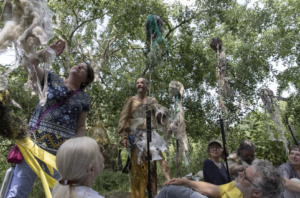 Seven people, most sat on the floor outdoors surrounded by green trees looking at materials attached to stakes.
